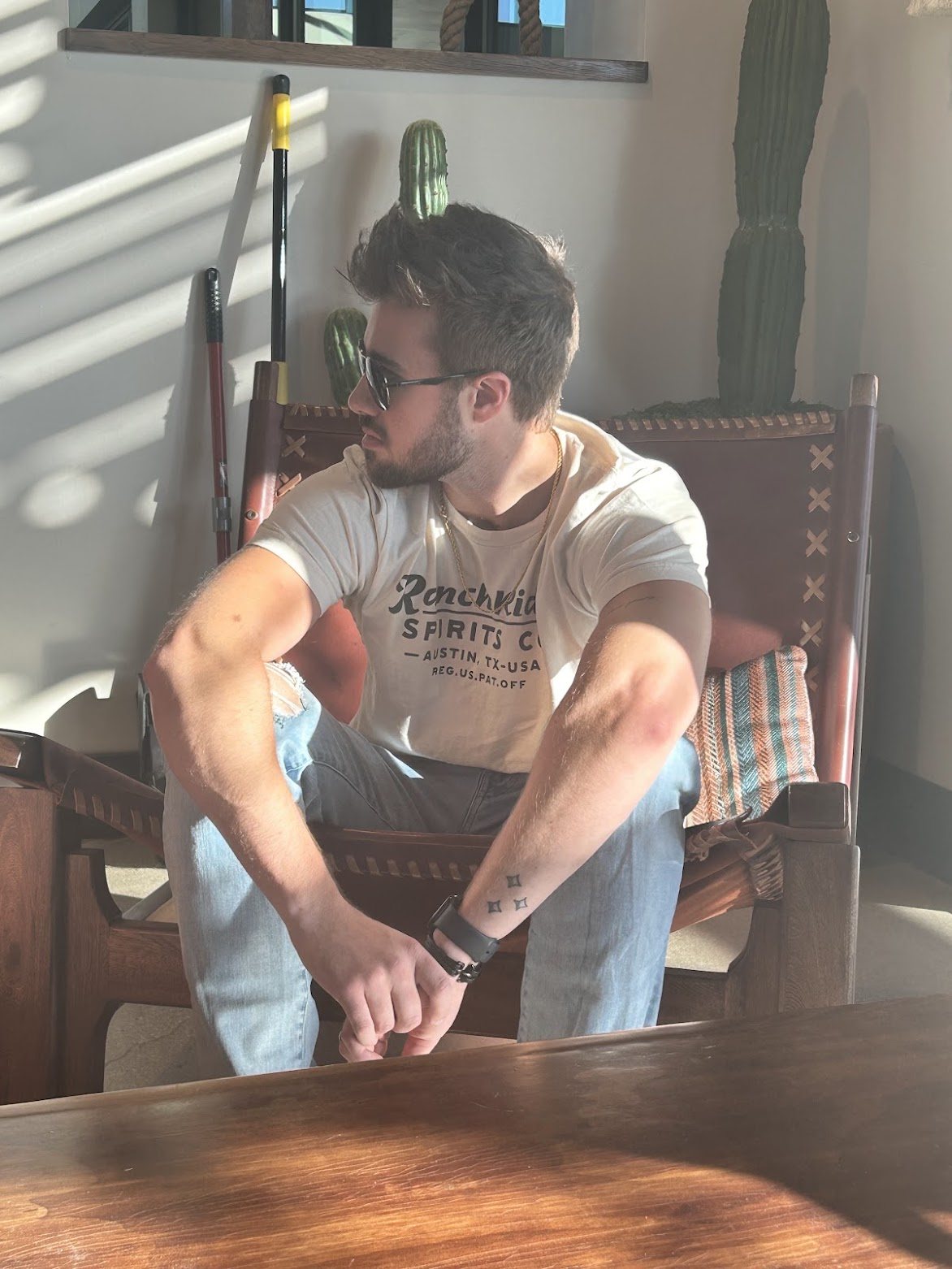 DJ Von Frank sitting in a leather and wood chair in profile, wearing sunglasses and a plain t-shirt, in a sunlit room with tall cacti behind him.