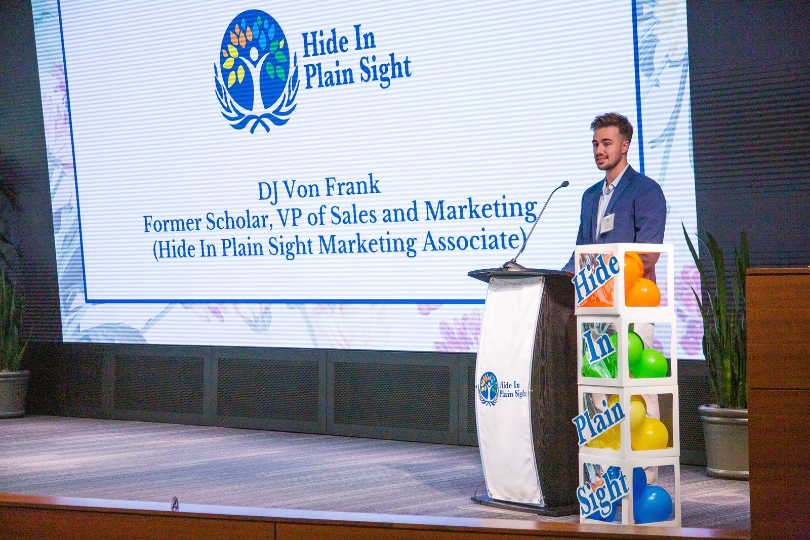 DJ Von Frank standing at a lectern beside a projected slide reading Hide In Plain Sight, which names him as a former scholar of the program.